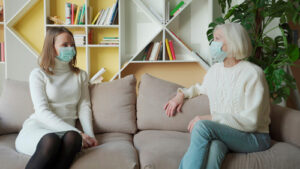 two woman sitting on couch talking and wearing masks